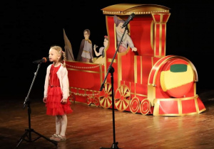 Na zdjęciu widać dziewczynkę stojącą przy mikrofonie, na drugim planie widać czerwoną lokomotywę wykonaną z papieru.